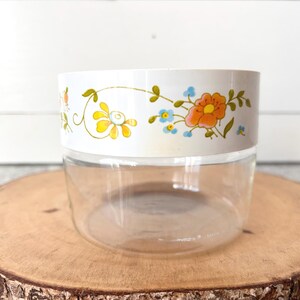 May include: A clear glass jar with a white band featuring a floral design in orange, yellow, and blue. The jar sits on a round wooden slice, showcasing a vintage aesthetic. Ideal for kitchen storage.