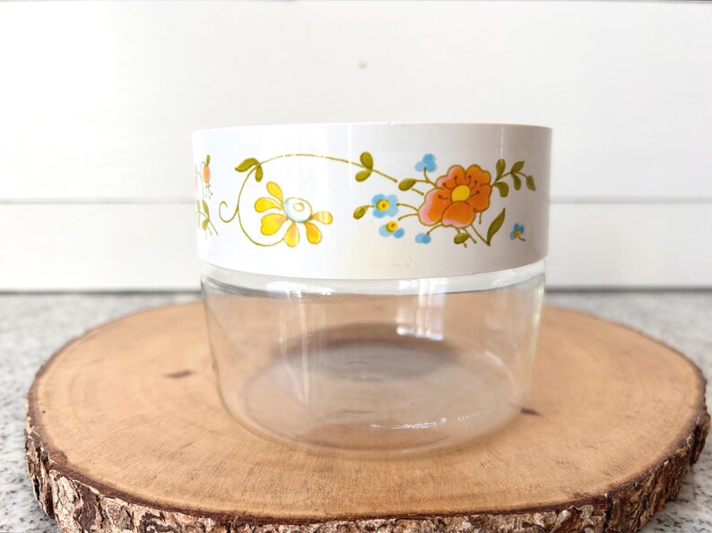 May include: A clear glass jar with a white band featuring a floral design in orange, yellow, and blue. The jar sits on a round wooden slice, showcasing a vintage aesthetic. Ideal for kitchen storage.