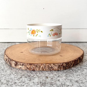 May include: A clear, round container with a white band featuring floral designs in orange, pink, and blue. The container sits on a round, wooden slice with a natural bark edge. The background is a white wall.