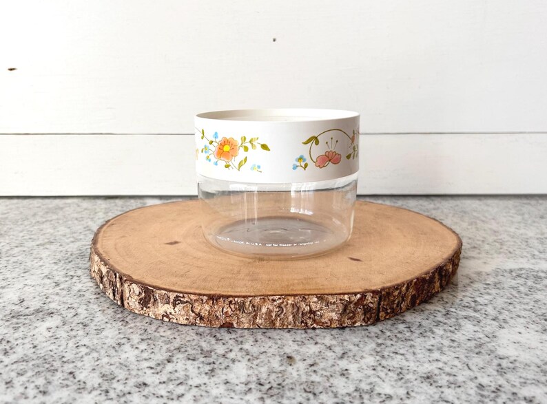 May include: A clear, round container with a white band featuring floral designs in orange, pink, and blue. The container sits on a round, wooden slice with a natural bark edge. The background is a white wall.