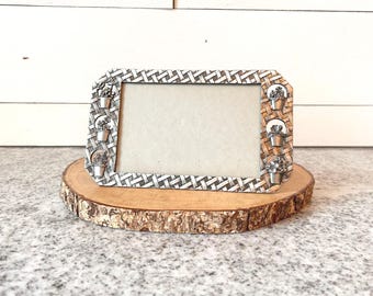 May include: Silver-toned picture frame with a woven pattern and small flower pot accents. The frame is displayed on a round, natural wood slice, creating a rustic aesthetic. The background is a white wooden wall.