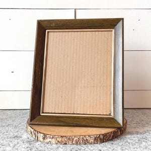 May include: A rectangular picture frame with a dark green wooden frame and a cardboard backing. The frame is displayed on a round wooden base with a natural bark edge. The background is a white wooden wall.