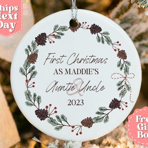 May include: White ceramic Christmas ornament with a green and brown pine bough wreath design. The ornament has the text "First Christmas as Maddie's Auntie & Uncle 2023".