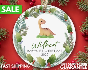 May include: A white round Christmas ornament with a green watercolor wreath, a brown dinosaur illustration, and the text "Wilbert Baby's 1st Christmas 2022".