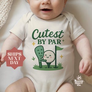 May include: A cream-colored baby onesie with the text "Cutest BY PAR" in green. The graphic features a golf club hugging a golf ball, with a green flag and yellow stars. The Sunny Heart Co. logo is in the bottom right.