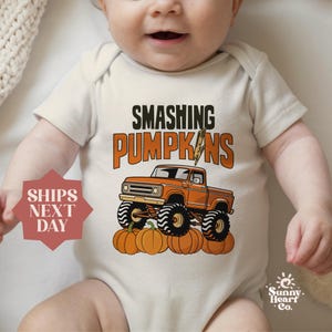 May include: Cream-colored baby onesie with the text "SMASHING PUMPKINS" in black and orange. An orange monster truck graphic is featured above a row of pumpkins. The onesie has short sleeves.