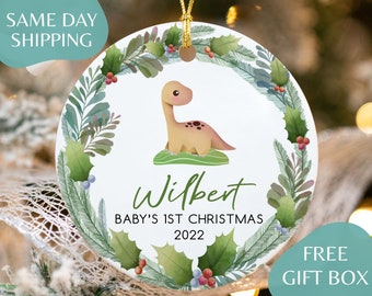 May include: A white ceramic Christmas ornament with a green holly wreath border. A cartoon dinosaur is in the center with the name "Wilbert" and the text "Baby's 1st Christmas 2022".