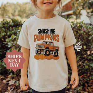 May include: Beige t-shirt featuring the text "Smashing Pumpkins" above an orange monster truck design. The truck is driving over pumpkins. The shirt also has the Sunny Heart Co. logo.