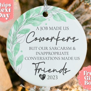May include: White round ceramic ornament with green watercolor leaves and the text "A job made us coworkers but our sarcasm & inappropriate conversations made us friends 2023".