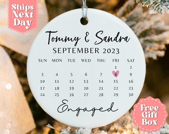 May include: White ceramic ornament with a calendar design featuring the month of September 2023. The date 8 is highlighted with a pink heart. The text "Tommy & Sandra Engaged" is written on the ornament.