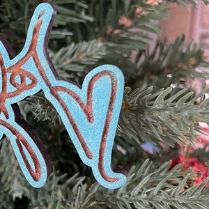 May include: A teal and brown wooden Christmas ornament in the shape of cursive letters, hanging from a white string. The ornament is layered, with a teal center and brown outline. The ornament is hanging on a Christmas tree.