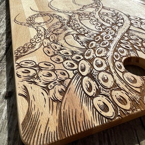 Puede incluir: Una tabla de cortar de madera con un diseño de un pulpo grabado con láser. El pulpo está detallado con tentáculos y ventosas. La tabla tiene un agujero en el centro para colgar.