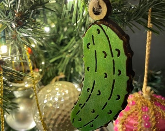 May include: A green wooden pickle ornament hanging on a Christmas tree. The ornament is shaped like a pickle and has a brown wooden string attached to it.