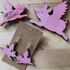 May include: A pair of pink pig earrings with wings. The earrings are on a brown card with the text "All things are possible".