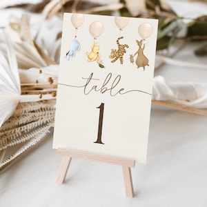 Puede incluir: Número de mesa uno con un tema de Winnie the Pooh. La tarjeta es beige con texto marrón e ilustraciones de personajes de Winnie the Pooh sosteniendo globos. La tarjeta está en un pequeño caballete de madera.