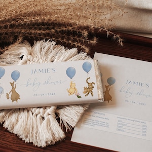 May include: A white chocolate bar wrapper with a blue and yellow Winnie the Pooh baby shower theme. The wrapper features Winnie the Pooh, Piglet, Tigger, and Eeyore holding blue balloons. The text on the wrapper reads "JAMIE'S baby shower 05 | 04 | 2023".
