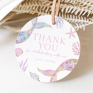 May include: A round white tag with a mermaid theme. The tag features two mermaid tails, seashells, and the text "THANK YOU" in pink. The tag is tied with a leather string.