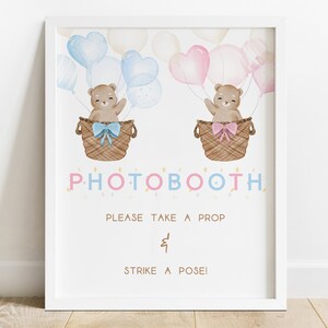May include: A white photo booth sign with a pink and blue color scheme. Two teddy bears are in hot air balloons with blue and pink balloons. The sign says "Photo Booth" with stars and the text "Please take a prop & strike a pose!"