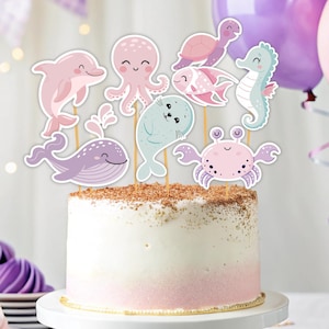 May include: A pink and purple cake decorated with sea animal cake toppers. The toppers include a whale, a crab, a seahorse, a seal, an octopus, a fish, and a turtle.