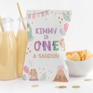 Puede incluir: Una bolsa de fiesta blanca con el texto "Kimmy is One A-Saurus!" en morado y verde. La bolsa presenta ilustraciones de dinosaurios, globos y un volcán. También se ven dos botellas de vidrio con pajitas y un cuenco de patatas fritas.