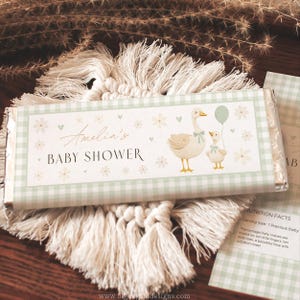 May include: A baby shower candy bar with a light green gingham pattern. The label features the text "Amelia's BABY SHOWER" and illustrations of a goose and gosling with a balloon. The bar rests on a white macrame coaster.