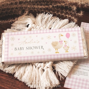 May include: A chocolate bar wrapper with a pink gingham border and the text "Amelia's BABY SHOWER." It features illustrations of a mother goose and a baby goose holding a pink balloon, with floral and heart accents. The wrapper is on a macrame coaster.