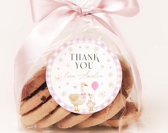 Könnte beinhalten: Ein klarer Zellophanbeutel, gebunden mit einem rosa Band, gefüllt mit Keksen. Ein runder Aufkleber mit einem rosa Vichy-Muster trägt die Aufschrift "THANK YOU love Amelia" mit einer Gänse- und Gänseblümchen-Illustration.