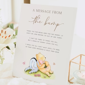 Op de afbeelding: Een witte kaart met gouden letters die "Een boodschap van de bult" zegt. De kaart heeft een boodschap over de komst van de baby en bevat een cartoonillustratie van Winnie de Poeh en Biggetje.