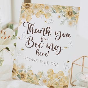 May include: A white sign with a yellow and brown floral design featuring sunflowers and bees. The sign says "Thank you for Bee-ing here! Please take one."