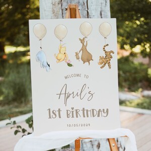 K&ouml;nnte beinhalten: Ein Winnie-the-Pooh-Geburtstagsschild mit f&uuml;nf gelben Ballons, einem blauen Esel, einem gelben B&auml;ren, einem braunen K&auml;nguru und einem orangefarbenen Tiger. Das Schild lautet "Willkommen zum 1. Geburtstag von April 10/05/2024".