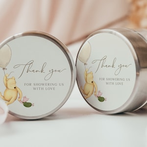 May include: Two round favor boxes with Winnie the Pooh and Piglet holding balloons. The boxes have a white background with brown text that says "Thank you for showering us with love".