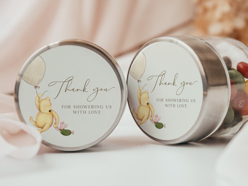 May include: Two round favor boxes with Winnie the Pooh and Piglet holding balloons. The boxes have a white background with brown text that says "Thank you for showering us with love".