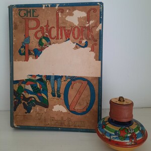 May include: An antique copy of "The Patchwork Girl of Oz" by L. Frank Baum, with a colorful illustration. Beside it is a vintage spinning top with a wooden handle and a painted design featuring circus performers and a red star.