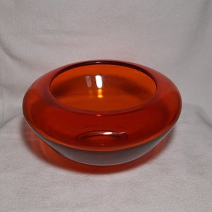 May include: A round, orange glass bowl with a wide opening. The bowl has a smooth, glossy surface and a dark base. The interior of the bowl reflects light, creating a warm glow. The bowl is likely used for decorative purposes.