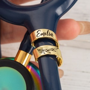 Op de afbeelding: Twee gouden stethoscoopbedels. De bovenste bedel is gegraveerd met de naam "Emelia". De onderste bedel heeft een delicaat bladontwerp.