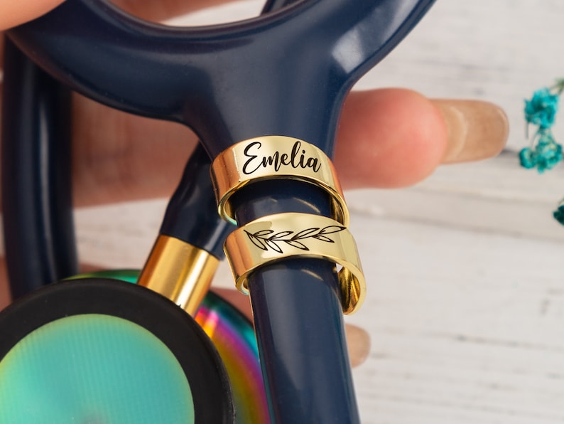 Op de afbeelding: Twee gouden stethoscoopbedels. De bovenste bedel is gegraveerd met de naam "Emelia". De onderste bedel heeft een delicaat bladontwerp.