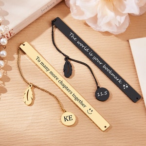 May include: Two gold and black metal bookmarks with chains and charms. The gold bookmark has the text "To many more chapters together" and a smiley face. The black bookmark has the text "The world is your bookmark" and a star. Both bookmarks have a feather charm.