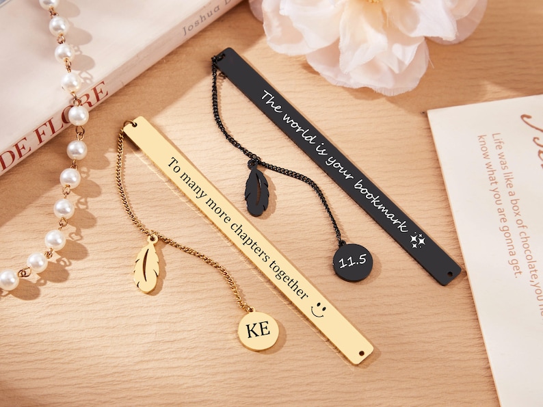 May include: Two gold and black metal bookmarks with chains and charms. The gold bookmark has the text "To many more chapters together" and a smiley face. The black bookmark has the text "The world is your bookmark" and a star. Both bookmarks have a feather charm.
