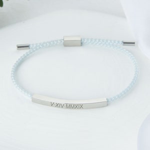 May include: A light blue braided bracelet with a silver-colored rectangular bar engraved with Roman numerals. The bracelet has a sliding knot closure and silver-colored end caps. The bracelet is a piece of jewelry.