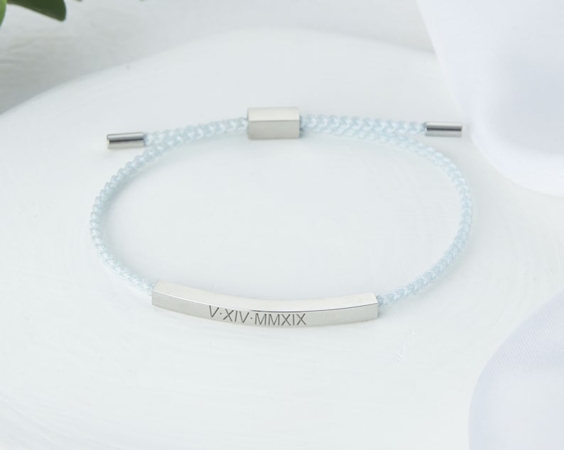 May include: A light blue braided bracelet with a silver-colored rectangular bar engraved with Roman numerals. The bracelet has a sliding knot closure and silver-colored end caps. The bracelet is a piece of jewelry.