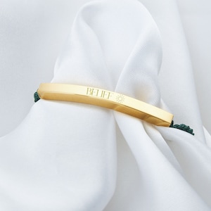 May include: A gold-colored bracelet with the word "BELIEF" and a sun symbol engraved on it. The bracelet is set against a white background and has dark green cords on each side.