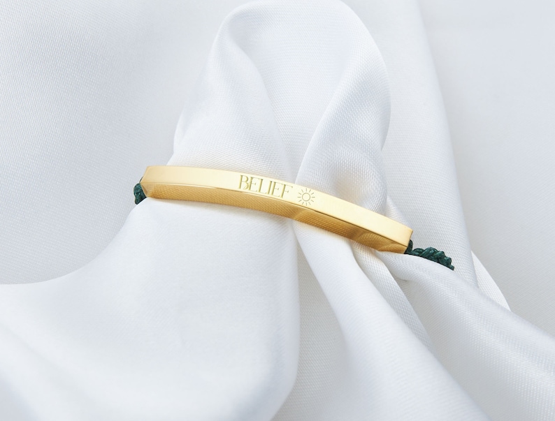 May include: A gold-colored bracelet with the word "BELIEF" and a sun symbol engraved on it. The bracelet is set against a white background and has dark green cords on each side.