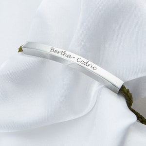 May include: A silver bar bracelet with the names "Bertha" and "Cedric" engraved on it. The bracelet is attached to a green cord. The background is a white, silky fabric.