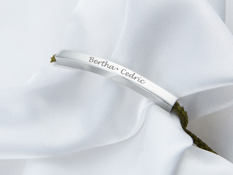 May include: A silver bar bracelet with the names "Bertha" and "Cedric" engraved on it. The bracelet is attached to a green cord. The background is a white, silky fabric.