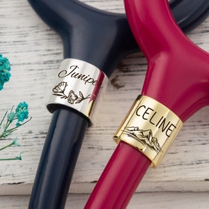 Op de afbeelding: Twee gepersonaliseerde stethoscoop-naambordjes. Een bordje is zilverkleurig met de naam "Junie" en een bloemmotief. Het andere bordje is goudkleurig met de naam "CELINE" en een bergmotief.