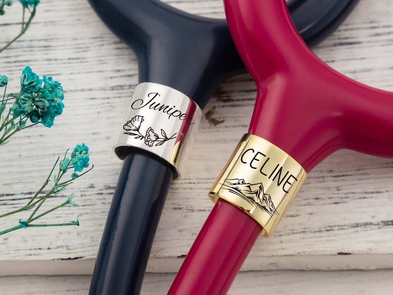 Op de afbeelding: Twee gepersonaliseerde stethoscoop-naambordjes. Een bordje is zilverkleurig met de naam "Junie" en een bloemmotief. Het andere bordje is goudkleurig met de naam "CELINE" en een bergmotief.