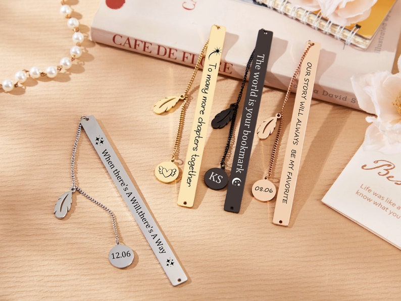 May include: Five gold, silver, and black metal bookmarks with engraved text and a dangling feather charm. The bookmarks have a chain attached to a round charm with a date engraved on it. The text on the bookmarks reads: "The world is your bookmark", "To many more chapters together", "When there's A Will, there's A Way", "Our story will always be my favorite", and "KS".