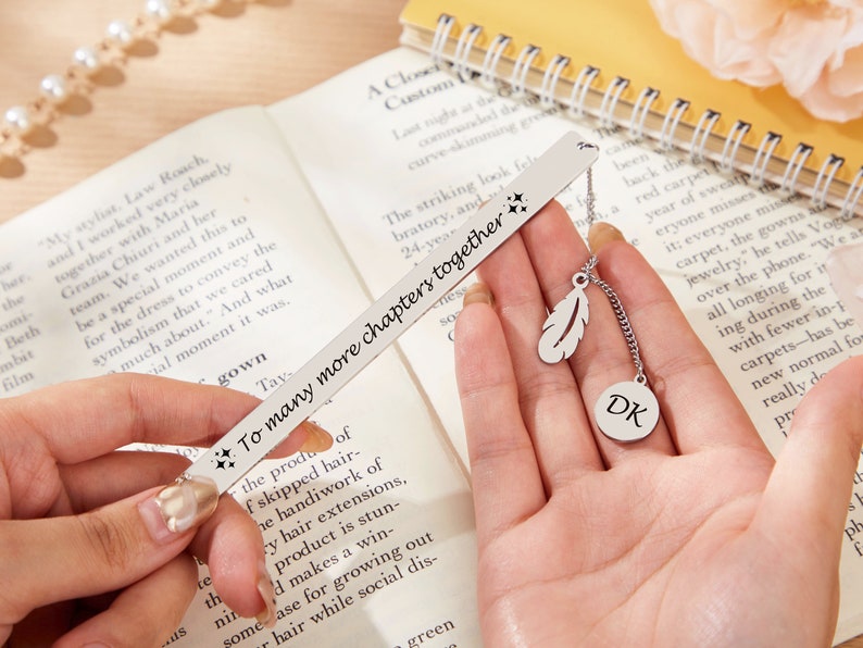 May include: Silver bookmark with a feather charm and a round charm with the initials "DK" engraved. The bookmark has the text "To many more chapters together" engraved on it.