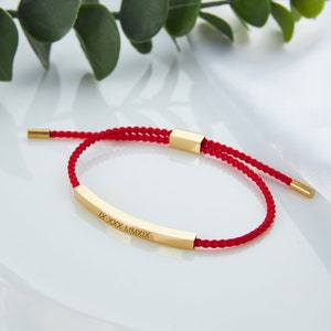May include: A red braided bracelet with a gold-colored rectangular bar. The bar is engraved with Roman numerals. The bracelet has adjustable sliding knots and gold-colored end caps. The background is white.