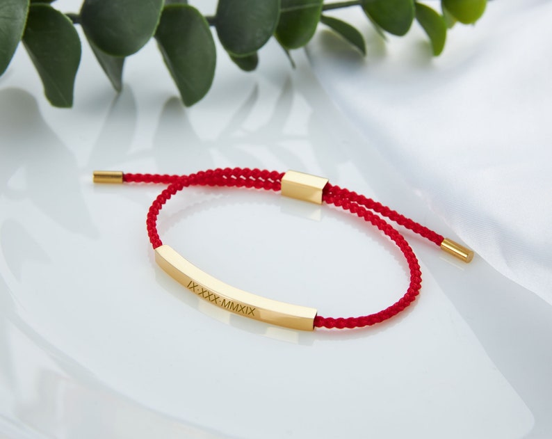 May include: A red braided bracelet with a gold-colored rectangular bar. The bar is engraved with Roman numerals. The bracelet has adjustable sliding knots and gold-colored end caps. The background is white.